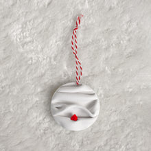 Load image into Gallery viewer, made from polymer clay, its a flat circle ornament with speckles white folds made to look like a snowy landscape, with a red house nestled on a hill, hung with a red and white string
