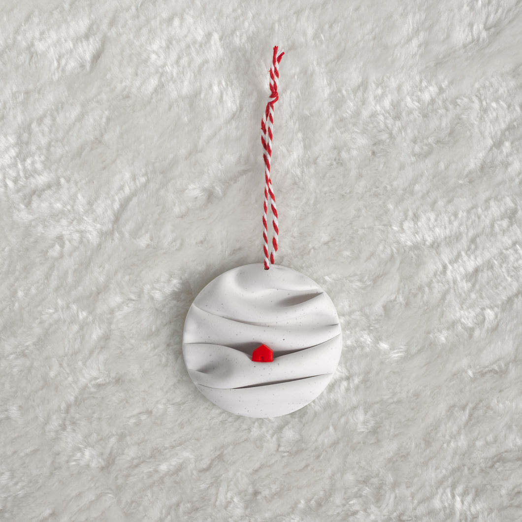 made from polymer clay, its a flat circle ornament with speckles white folds made to look like a snowy landscape, with a red house nestled on a hill, hung with a red and white string