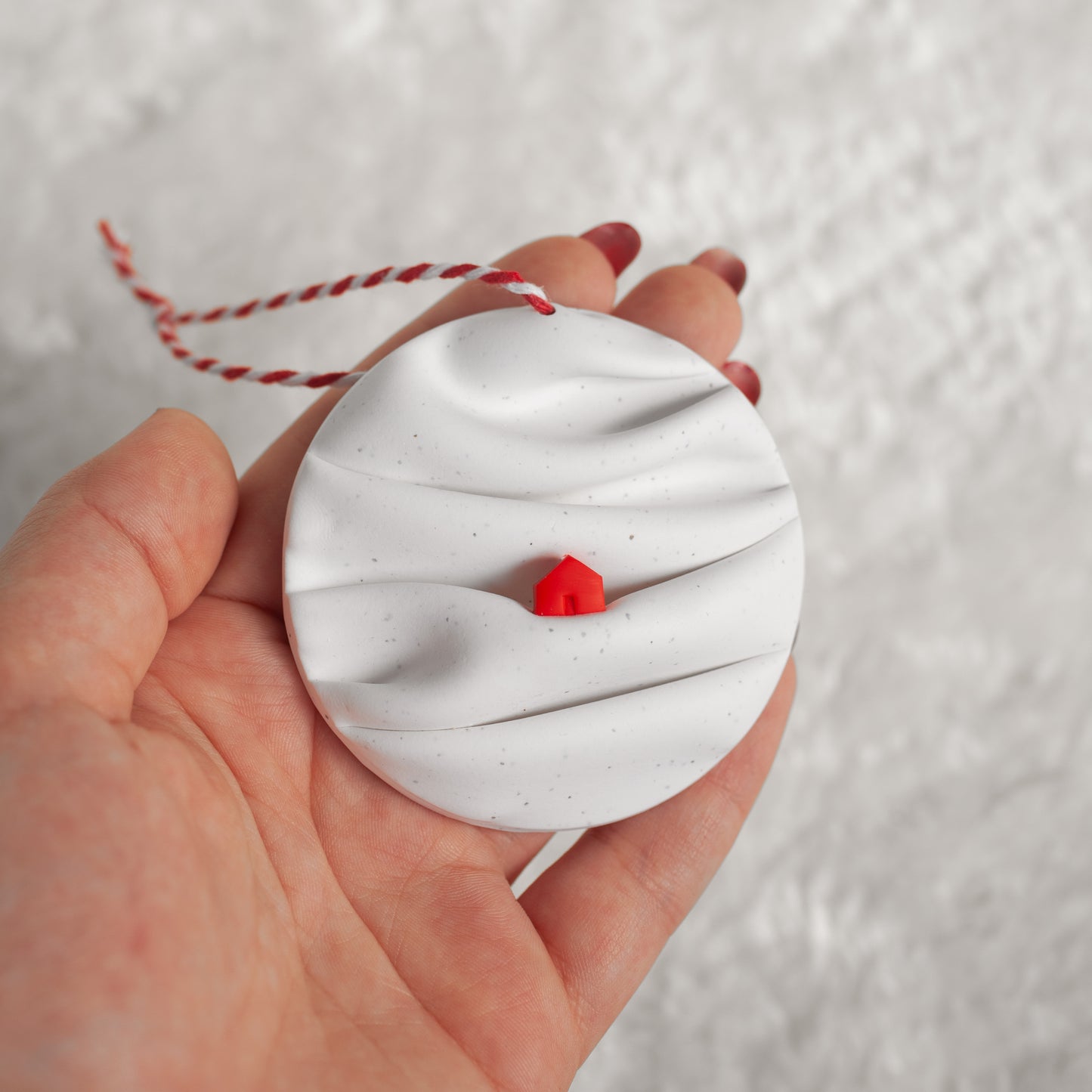 made from polymer clay, its a flat circle ornament with speckles white folds made to look like a snowy landscape, with a red house nestled on a hill, hung with a red and white string