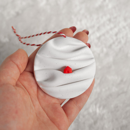 made from polymer clay, its a flat circle ornament with speckles white folds made to look like a snowy landscape, with a red house nestled on a hill, hung with a red and white string